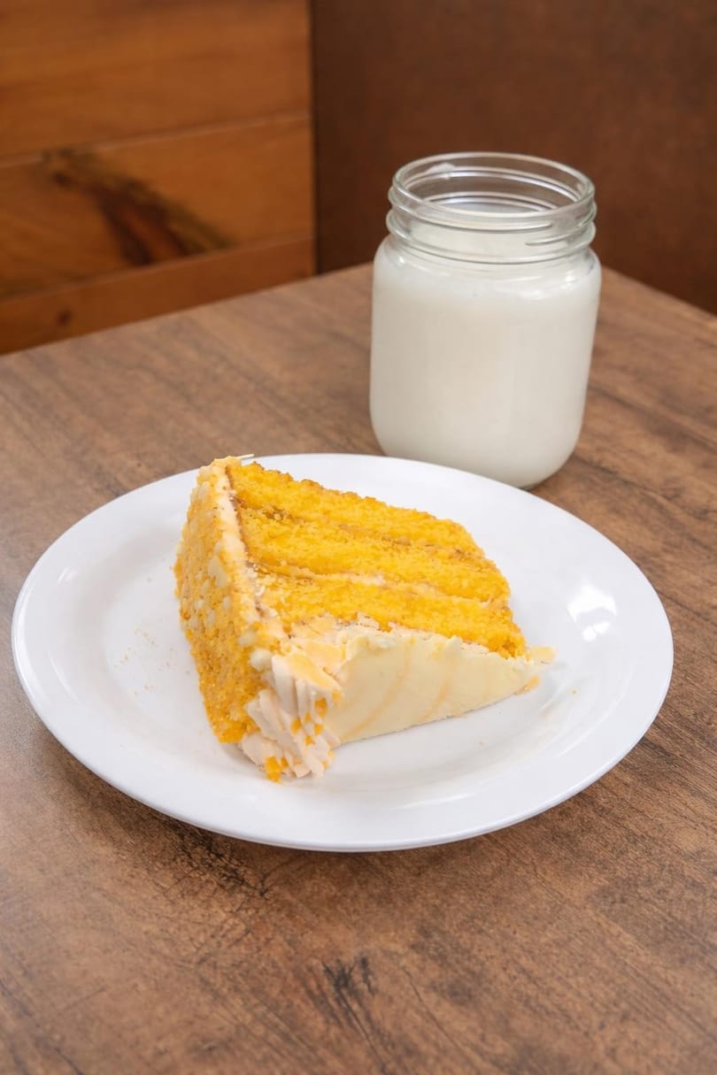 A slice of yellow cake with white frosting and a glass of milk on a wooden table.