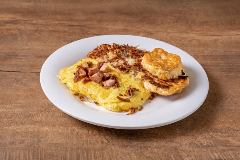 Breakfast plate with a ham and bacon omelet, hash browns, and a biscuit on a white plate.