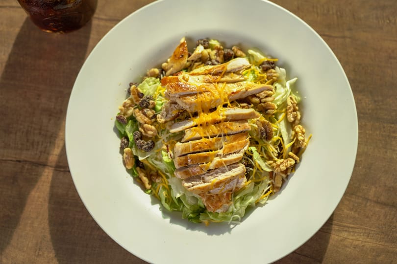 Grilled chicken salad with lettuce, walnuts, raisins, and shredded cheese on a white plate.