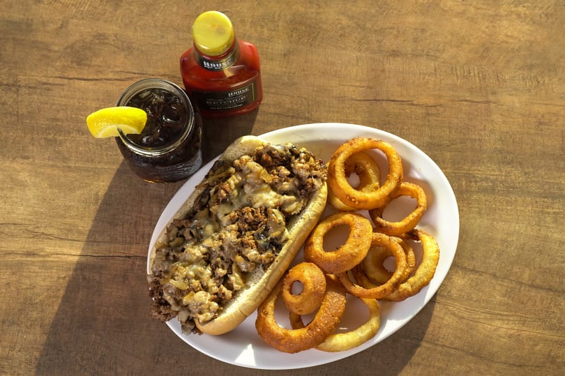 Cheesesteak sandwich with mushrooms and onions, onion rings, soda with lemon, and ketchup on a wooden table.